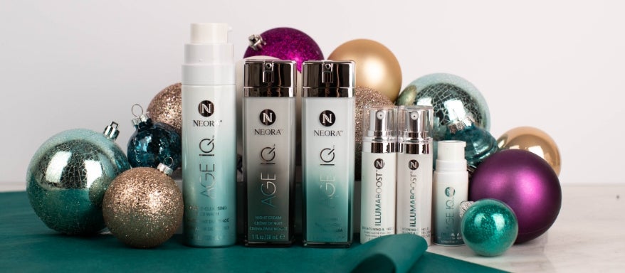 Image of the Age IQ skincare products, including Double Cleansing Face Wash, Night Cream, Day Cream, IllumaBoost Vitamin C Serum and Age IQ Eye Cream in front of vibrantly colored Christmas ornaments.