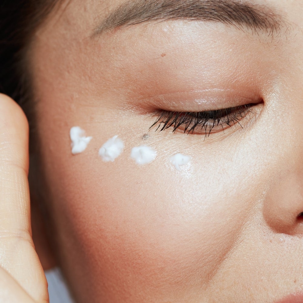 A closeup of a woman’s face with four dots of moisturizer under her right eye.