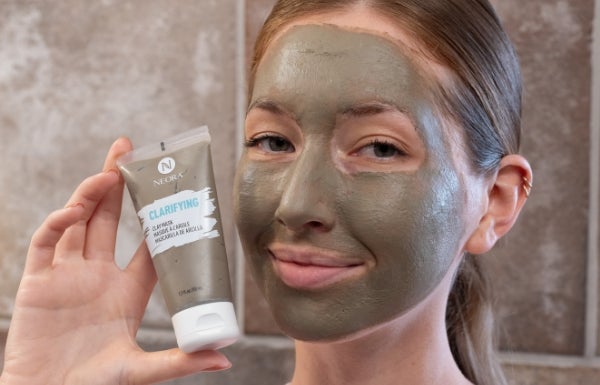 A close up of a woman’s face covered with Neora’s Clarifying Clay mask. She holds up a tube of this product next to her face.