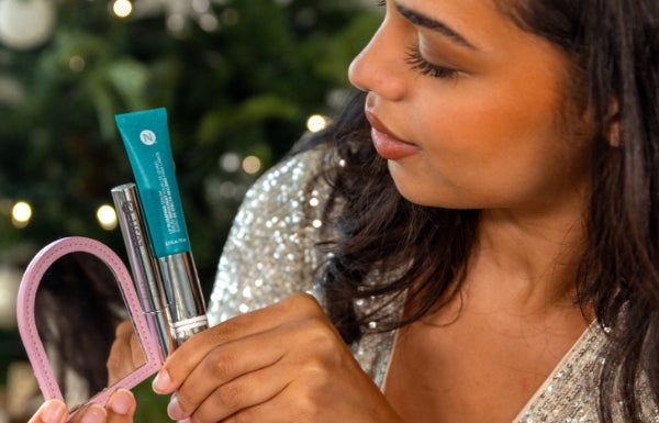 A woman holds up a tube of LashLush, Lip Pumping Serum and a pink Purse Mirror in front of a festive tree.