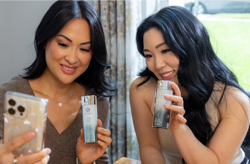 Two women take a selfie with Age IQ Day and Night Cream bottles.