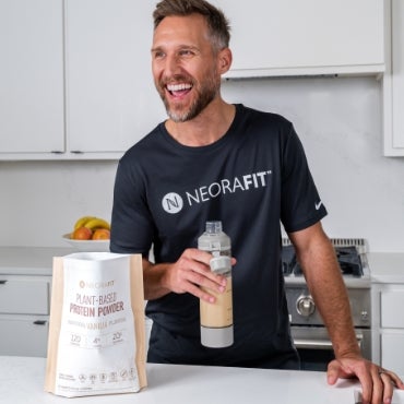 A man wearing a black NeoraFit shirt next to a kitchen counter holding up a glass of water with a bag of Neora's Plant-Based Protein Powder in front of him.