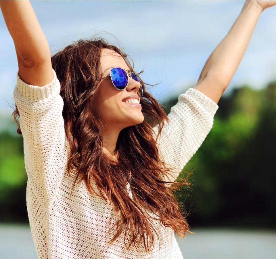 A woman wearing sun glasses and a white sweater embracing sunlight outside.