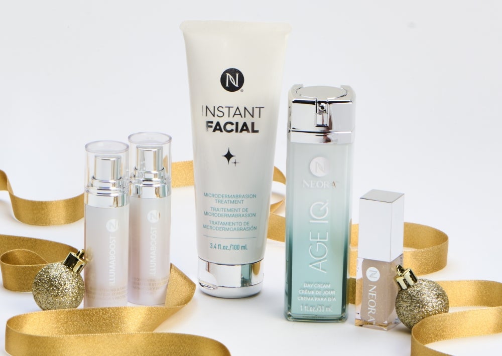 Los frascos de IllumaBoost, el tubo de Instant Facial de Neora, la crema de día Age IQ y un envase de aceite labial brillante de Neora están rodeados de un lazo dorado y adornos. El texto adjunto dice: ¡Regalo gratis! Y ahorra un 20 %.